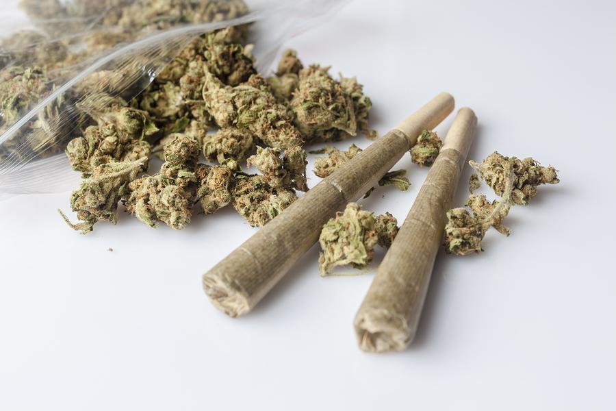 Pile of medical cannabis dried buds scattered from nylon package and two marijuana joints on white background from side