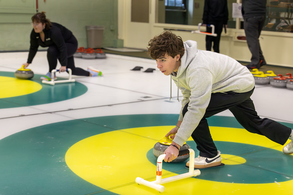 Nolan Suraci 26 delivers a curling stone, indicated by his lunge position with one hand on the handle of the stone and the other outstretched for balance. 